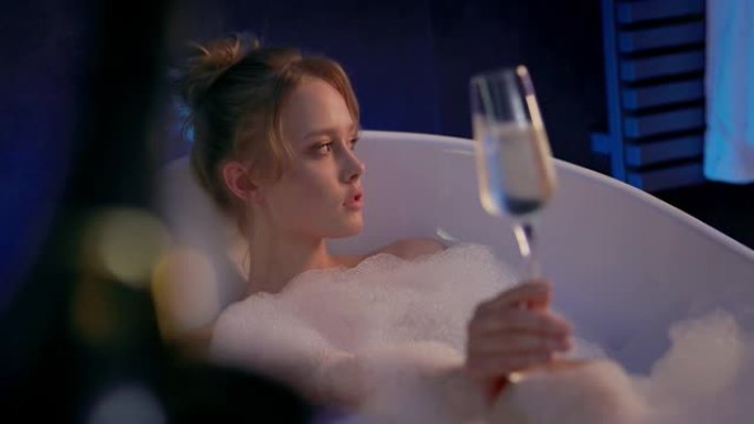 可爱的金发女郎在浴缸里喝一杯香槟。浴缸里有很多泡沫。女孩放松。