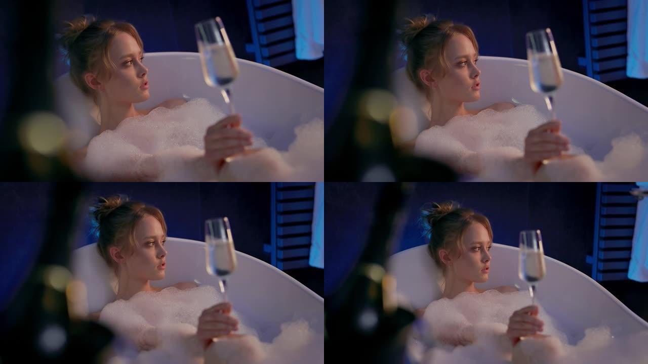 可爱的金发女郎在浴缸里喝一杯香槟。浴缸里有很多泡沫。女孩放松。