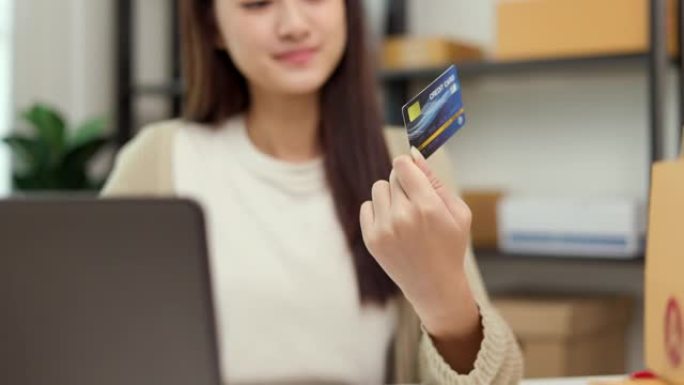 网上购物。年轻的亚洲妇女将信用卡的序列号输入到笔记本电脑上进行购物付款。在家在线送货购买商品。桌上有