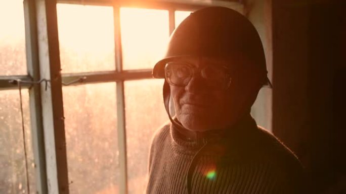 戴眼镜的老人在灰尘中捡起头盔，在房子窗户里戴上头盔