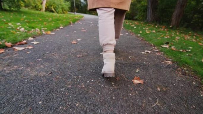一个女人在秋天的公园里沿着一条小路走，在框架中只能看到她的腿
