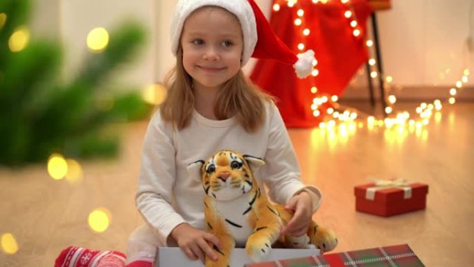 穿着红色圣诞老人帽子的快乐小女孩坐在打开的盒子和玩具老虎，看着相机，在圣诞树的背景上微笑。
