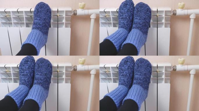 在寒冷的冬日，一名妇女穿着蓝色羊毛袜在散热器上温暖脚。中央供暖系统。寒冷季节昂贵的取暖费用