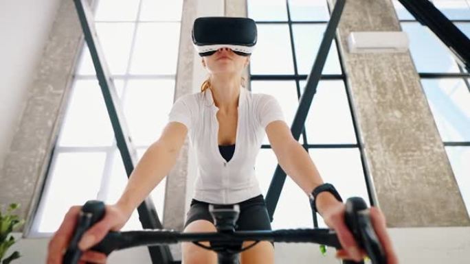 戴VR眼镜的年轻女性正在踩踏室内自行车教练。工作室公寓，厨房。在家进行日常训练。特写，慢动作，低角度