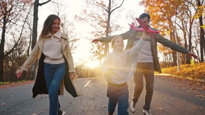 年轻的父母笑着，带着小女儿拿着红色的玩具飞机在马路上奔跑。秋公园。幸福的家庭。特写，慢动作。跟踪镜头