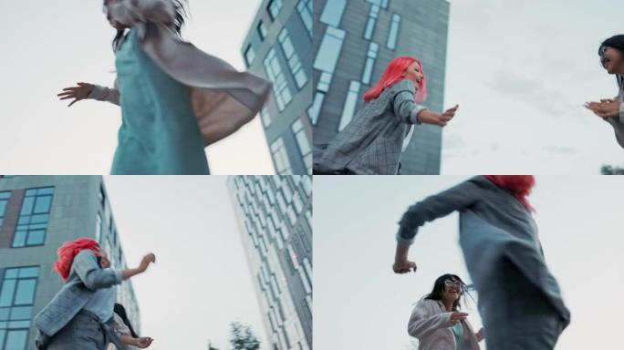 两个女孩穿着优雅的跳跃，在玻璃现代公司大楼前手拉手跳舞，同事的融合，晋升的喜悦，在工作中获得工作娱乐