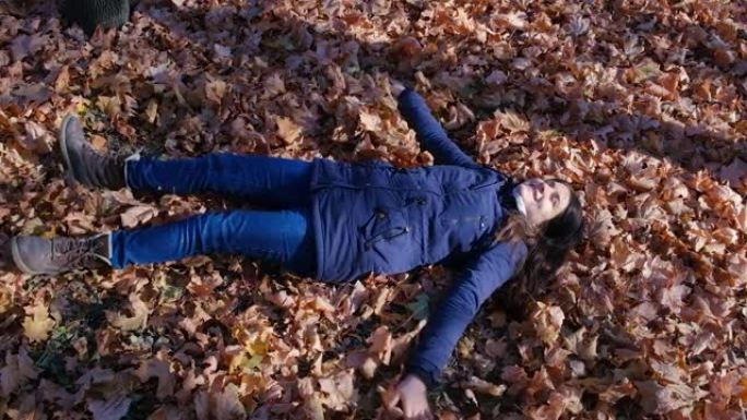 一个女人躺在秋天的森林里，躺在落叶上，举起双手。