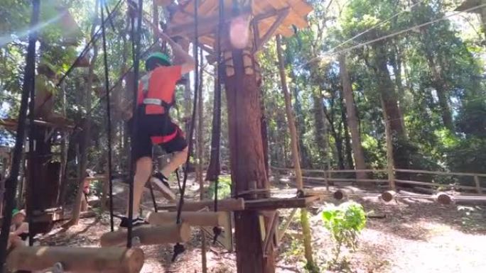 一个快乐的小男孩在少年儿童雨林冒险公园操场上平衡并走过一座绳索和悬挂原木的桥。
