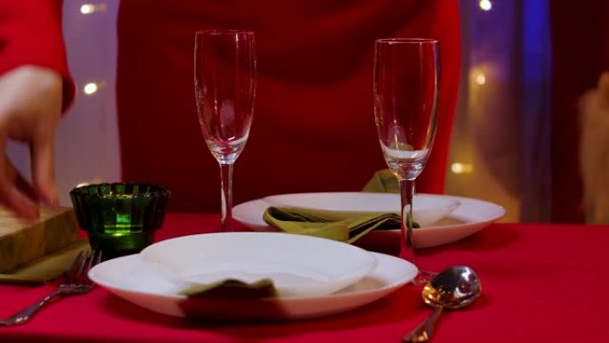 在节日的餐桌上，一位身着红色礼服的女士将餐叉和餐勺放在盛香槟的盘子和杯子旁边。家庭舒适的房间，装饰庆