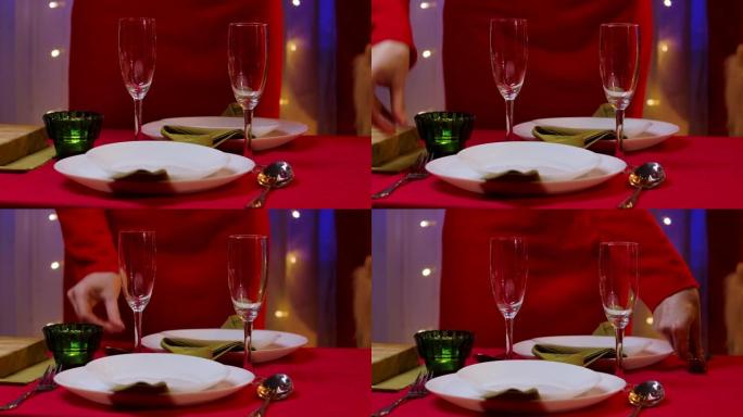 在节日的餐桌上，一位身着红色礼服的女士将餐叉和餐勺放在盛香槟的盘子和杯子旁边。家庭舒适的房间，装饰庆