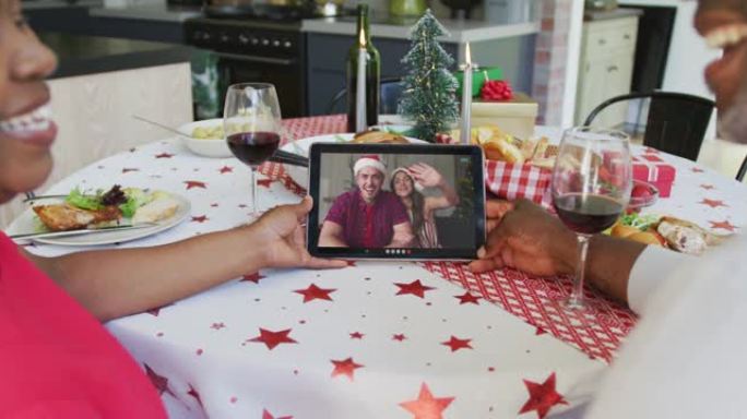 非裔美国人夫妇与葡萄酒使用平板电脑进行圣诞节视频通话与屏幕上的幸福夫妇