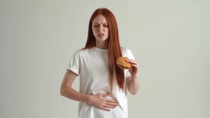 饥饿的年轻女子吃汉堡和胃痛，感觉恶心，食物中毒症状，胃炎的肖像。