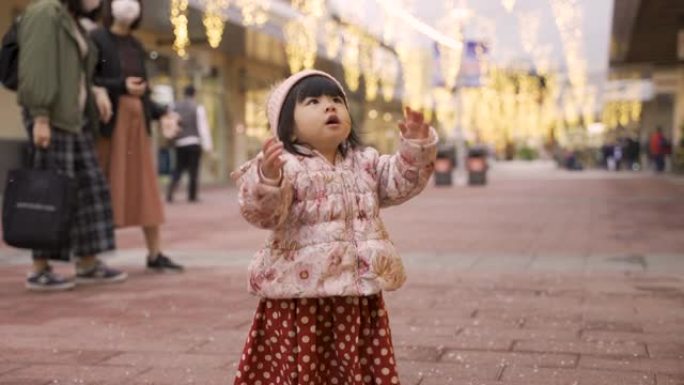 激动的中国女婴举起双手，看着天空中的飞雪，同时在圣诞节的白雪皑皑的街道上玩得开心