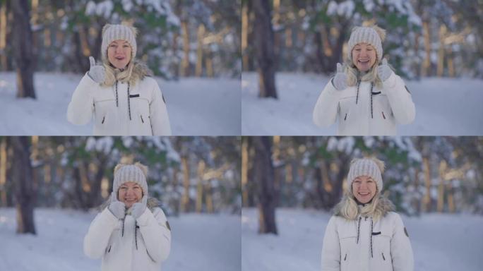 穿着温暖的冬装的可爱女孩的肖像微笑着竖起大拇指，手势像。年轻女子在白雪皑皑的森林模糊背景下在户外摆姿