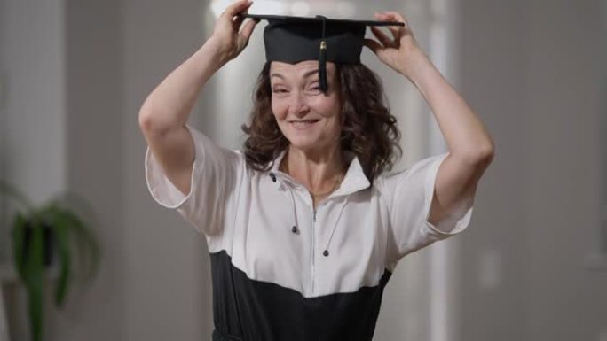 前视图聪明骄傲的成熟女人戴上毕业帽微笑着看着相机。自信的高加索老年毕业生在家里摆姿势的中拍肖像。动机