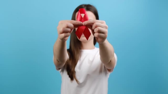 年轻女孩的选择性焦点手持红丝带向相机展示，蓝色背景上的模型。艾滋病治疗预防和保护，志愿者支持慈善事业