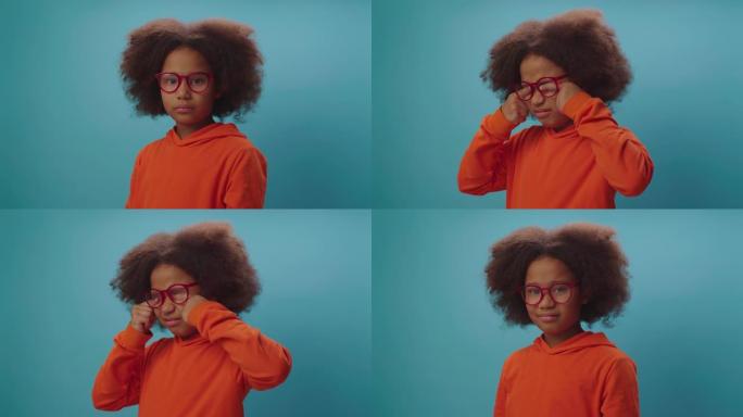 漂亮的黑人女孩厌倦了戴眼镜，站在蓝色背景上揉眼睛。戴护目镜有视力问题的孩子。