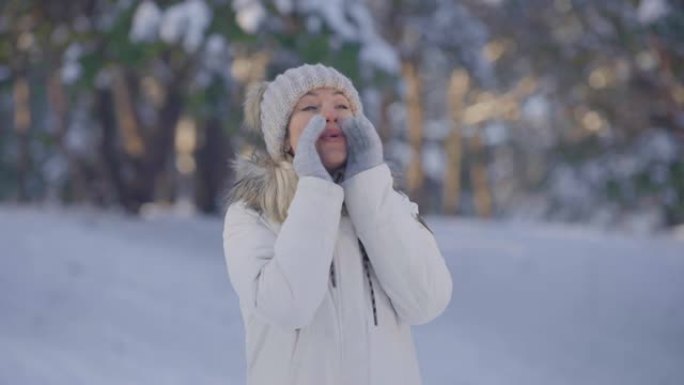 穿着暖和冬装的可爱女孩的肖像尖叫着叫人。年轻女子在白雪皑皑的森林模糊背景下在户外摆姿势。特写。慢动作