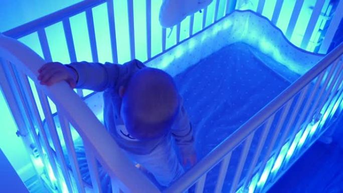 可爱的蹒跚学步的小孩晚上站在婴儿床里，快乐的男婴在婴儿床的灯光下发光夜色照明。黑暗房间里点燃的婴儿床