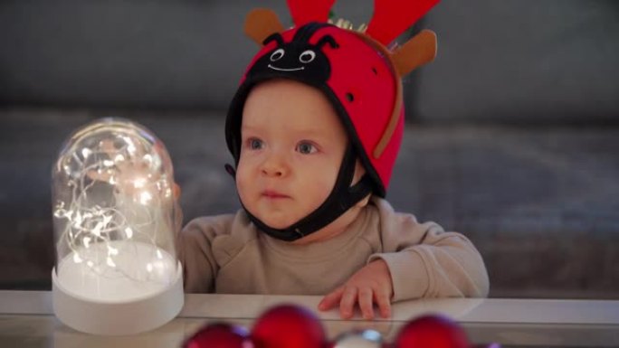 快乐的圣诞老人小帮手，红色驯鹿角，玩着圣诞装饰品，新年前夕拿着装饰照明的男婴。新年时间。假日季节。