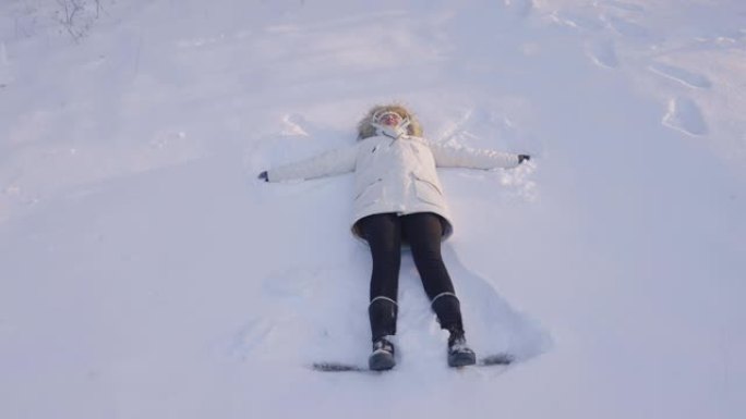 女孩在森林的一片空地上做了一个雪天使，从上面拍了一枪。快乐的年轻女子躺在雪地上移动手臂和腿。阳光明媚