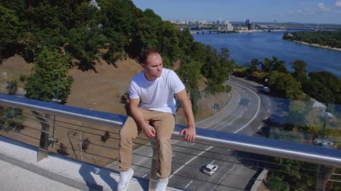 在城市的背景下，一个坐在玻璃桥栏杆上的家伙的跟踪镜头。高加索人坐在一根狭窄的金属管上。失去希望和自杀