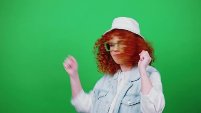 年轻的正面卷发红发女士戴着眼镜和巴拿马帽子走路跳舞，指着相机