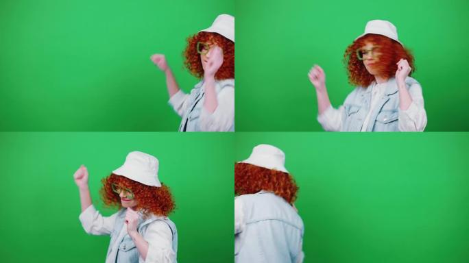 年轻的正面卷发红发女士戴着眼镜和巴拿马帽子走路跳舞，指着相机
