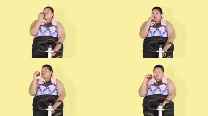 超重的女人在健身车上吃苹果