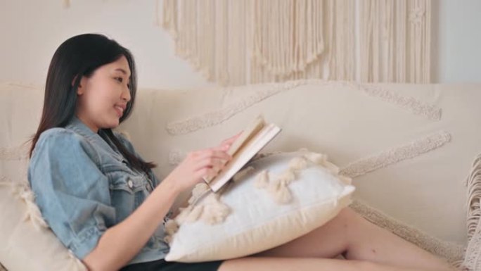 坐在沙发上靠在客厅的亚洲华裔妇女在深夜享受读书