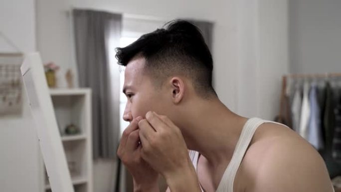侧视特写不安的亚洲年轻人担心面部皮肤问题正试图在早上在他的卧室里在镜子前除掉痤疮