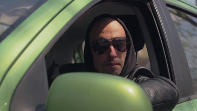 一个戴着兜帽和太阳镜的男人坐在汽车的方向盘后面，看着窗外