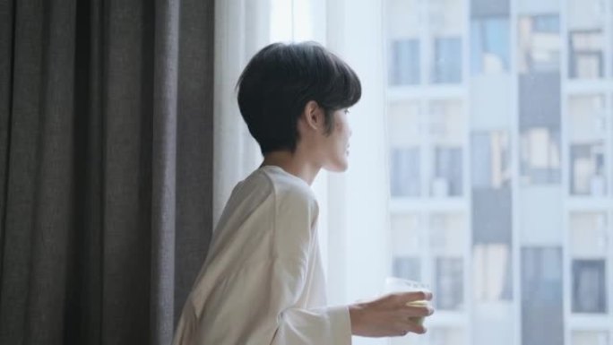 午餐休息时，亚洲妇女在家里工作时留着短发，拿着一杯冷饮走到窗前，从公寓内愉快而和平地看到风景