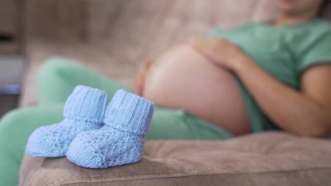 漂亮的针织袜子，适合未来的宝宝。准妈妈躺在沙发上的模糊背景上的新蓝色袜子。怀孕时间。