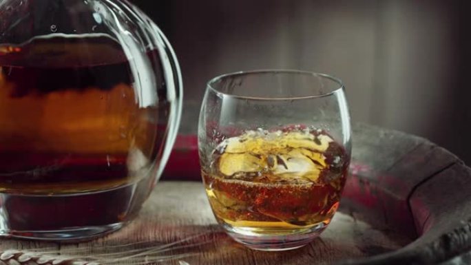 一块冰块慢慢落入老式威士忌桶顶部的一杯威士忌中。附近有一个装有饮料的decanter水器，背景为深棕