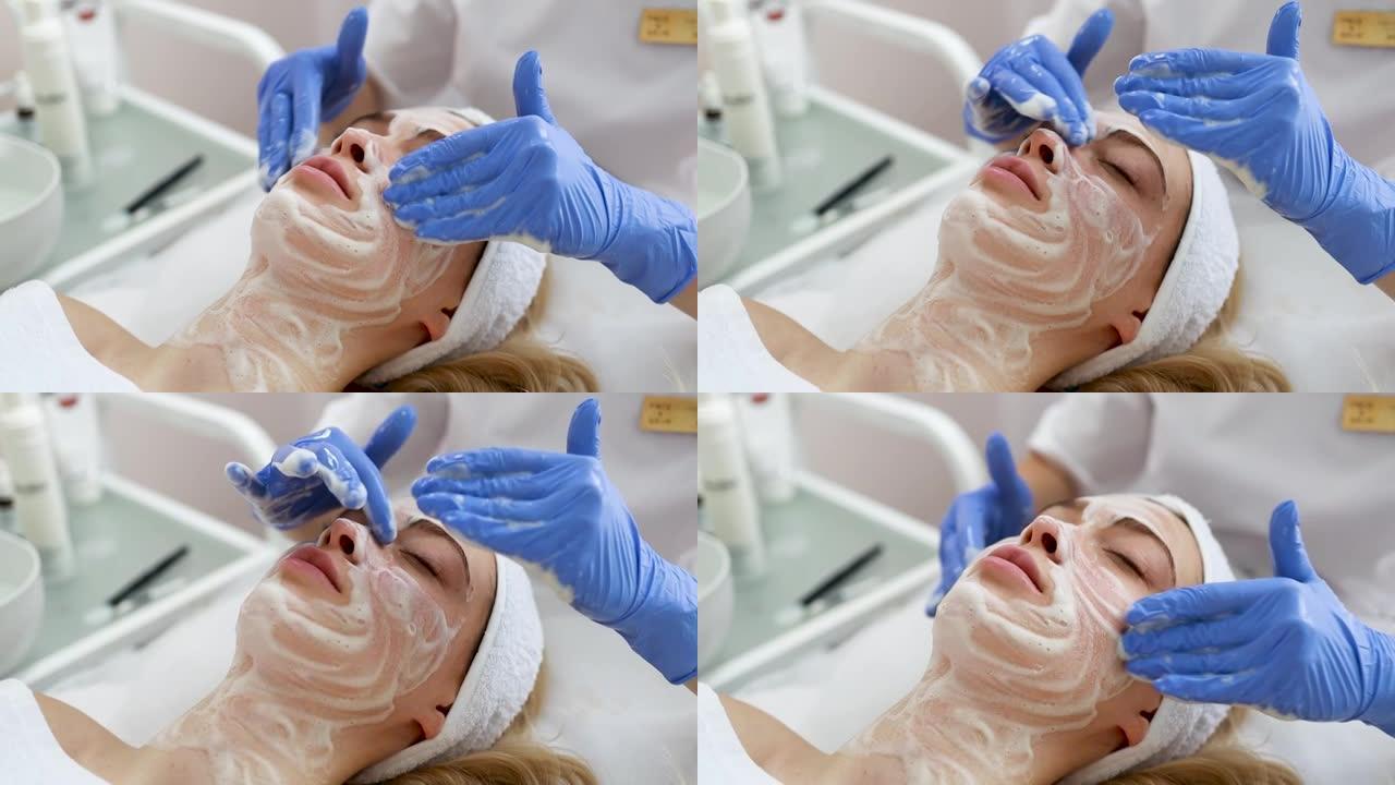 美容师医生正在在女人的脸上涂抹洁面泡沫。美容诊所的面部皮肤按摩