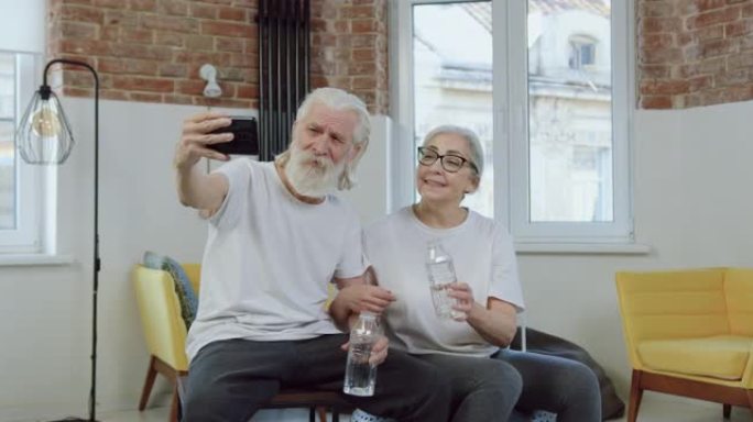 好看的微笑运动的老夫妇在家里锻炼时与朋友或亲戚进行视频通话时，用水碰碰瓶子，前视图