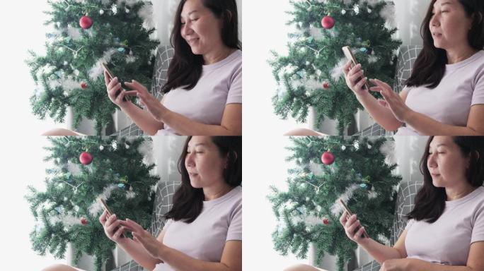 亚洲成年女性在家中使用智能手机附近的圣诞树装饰，生活方式概念。