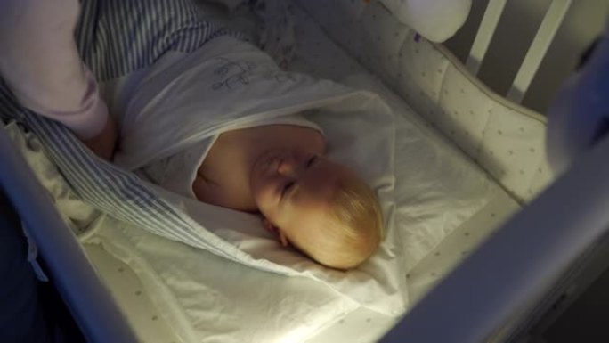 母亲襁褓中的婴儿睡在婴儿床里，面对襁褓中的9个月男婴在夜灯下。