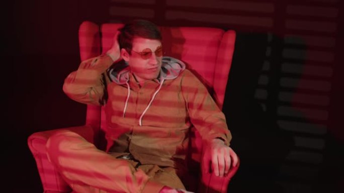 有思想的戴眼镜的男人坐在黑暗的房间里的扶手椅上，红色的光线和阴影的百叶窗。沉思的男性用手指数着，然后