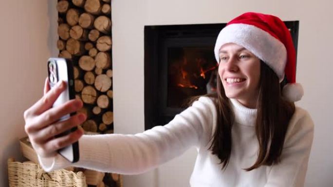 最美好的祝愿。快乐可爱的年轻女性视频博客作者坐在圣诞帽壁炉附近的温暖地板上，用电话迎接寒假的观众。千