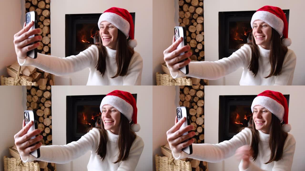 最美好的祝愿。快乐可爱的年轻女性视频博客作者坐在圣诞帽壁炉附近的温暖地板上，用电话迎接寒假的观众。千