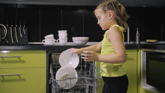 聪明的女孩学习使用洗碗机。时尚现代内置绿色黑色厨房电器。孩子正在放脏盘子