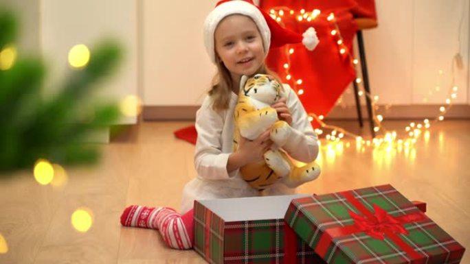 戴着红色圣诞老人帽子的快乐小孩在圣诞树旁边打开带有礼物玩具老虎的盒子。中国十二生肖新年的象征