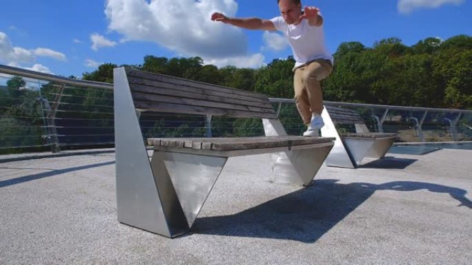 杂技演员在城市背景下在玻璃人行天桥的长凳上表演跑酷技巧的前视图。穿着米色裤子和白色t恤的高加索人翻滚