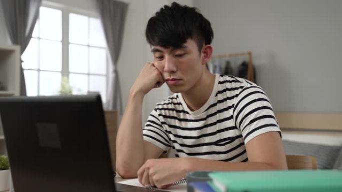 无聊的亚洲成年学生在家学习时，在锁定期间在卧室的计算机上学习在线课程时，正在支撑着脸并玩笔。