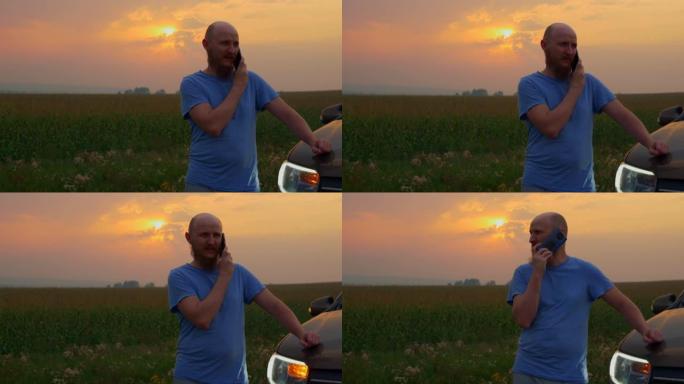 日落时分，一个留着胡须的秃头男人站在玉米田旁路边的一辆破车旁，打电话给技术服务或拖车。汽车在道路上出