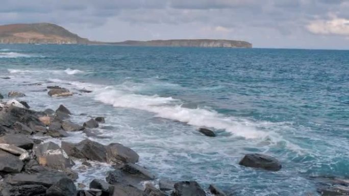 海浪溅落在蓝色海洋背景的岩石海岸上。波涛汹涌的岩石海岸线在暴风雨中席卷而来。海浪用泡沫在石头上破裂。