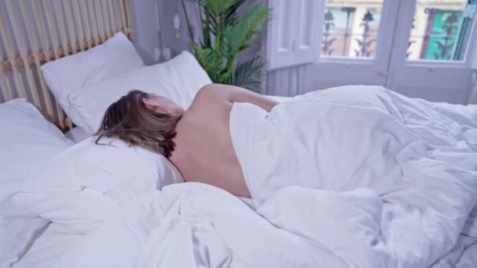 年轻女子在舒适的床上左侧卧。裸女在明亮舒适的卧室。假期早晨健康休息，不穿睡衣享受梦乡