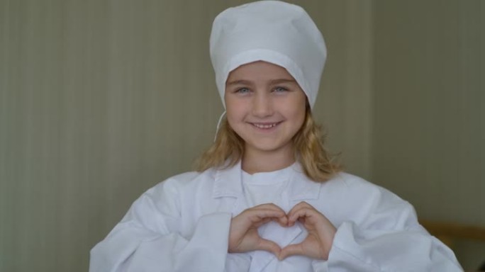 可爱的小女孩穿着医疗制服，用手做心脏标志。穿着医生制服和听诊器庆祝医生日的创意孩子微笑的肖像。心象征